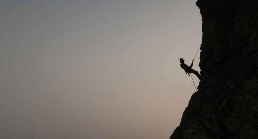 the silhouette of a rock climber resting midway up a rock wall